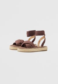 Sandale espadrile împletite, de culoare maro, cu bretele încrucișate și legături la glezne, având o platformă naturală din jute și talpă plată.
