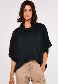 Poncho en tricot de couleur vert foncé avec un col drapé, présentant des manches larges et une coupe ample, associé à un pantalon à cordon léger beige.
