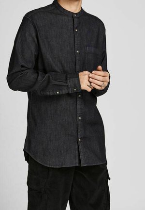 Homme portant une chemise en jean foncée à boutons avec un col mandarin et un pantalon cargo noir, les mains jointes à la taille.