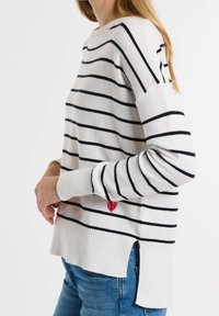 Gestreifter Pullover in Creme und Marineblau mit langen Ärmeln, seitlichen Schlitzen und einem roten Herzdetail nahe dem Saum. Weiche Stricktextur.