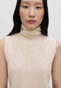 Beige mouwloze coltrui van zacht gebreid materiaal, met een hoge, opgerolde kraag en een gladde textuur. Minimalistisch ontwerp en aansluitende pasvorm.