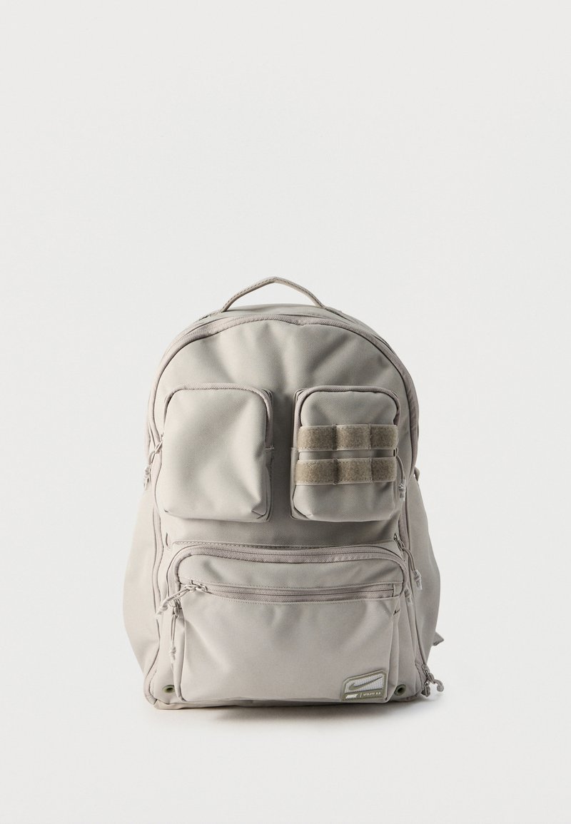 Ανοιχτό γκρι σακίδιο πλάτης με τρεις μπροστινές τσέπες με φερμουάρ, δύο μικρές επάνω τσέπες εκ των οποίων η μία έχει λουράκια με Velcro, και μια επάνω λαβή μεταφοράς.