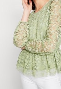 Blusa in pizzo verde chiaro con maniche a palloncino lunghe, vita arricciata, motivi floreali e sezioni trasparenti. Tessuto stratificato con dettagli delicati.