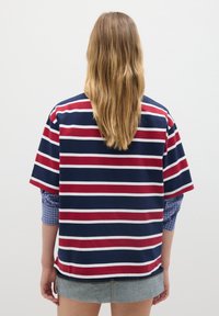 Camisa de manga corta a rayas en azul marino y blanco con acentos rojos, de corte relajado y dobladillo redondeado, superpuesta sobre una camisa de cuadros en azul.