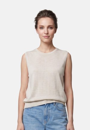 Young woman with brown hair tied back wears a sleeveless beige top and blue jeans, standing against a plain light background.