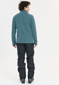 Teal pitkähihainen paita, jossa on korkea kaulus, yhdistetty mustiin lumihousuihin. Molemmilla on sileä pinta ja vartalonmyötäinen malli, joka on ihanteellinen ulkoilutoimintoihin.