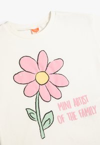 Witte T-shirt met een groot roze bloemenontwerp met een gele center en groene bladeren. De tekst luidt "MINI KUNSTENAAR VAN DE FAMILIE" in het roze.