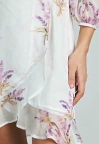 Leichtes, durchsichtiger weißes Kleid mit lavendelfarbenem Blumenmuster. Besitzt einen fließenden asymmetrischen Saum und Puffärmel, hergestellt aus einem weichen Stoff.
