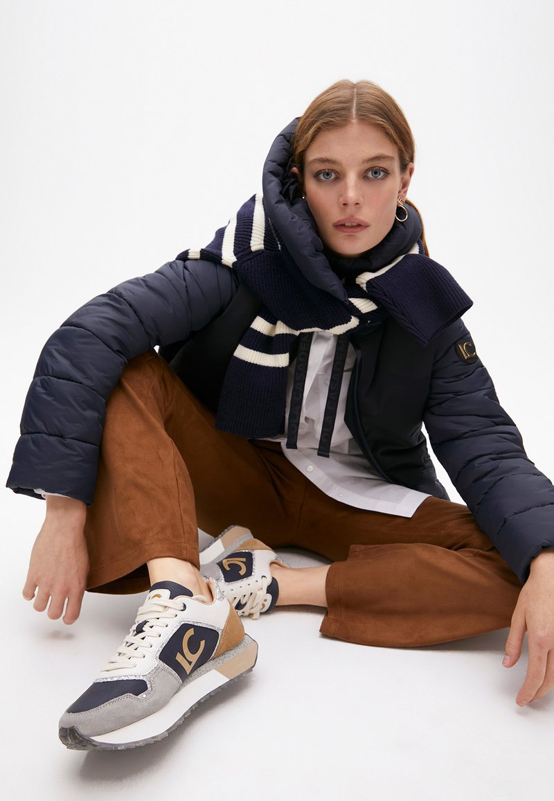 Giubbotto imbottito blu, camicia bianca, pantaloni a gamba larga marroni; sneakers blu e grigie con dettagli testurizzati e logo. Sciarpa con strisce blu e bianche.