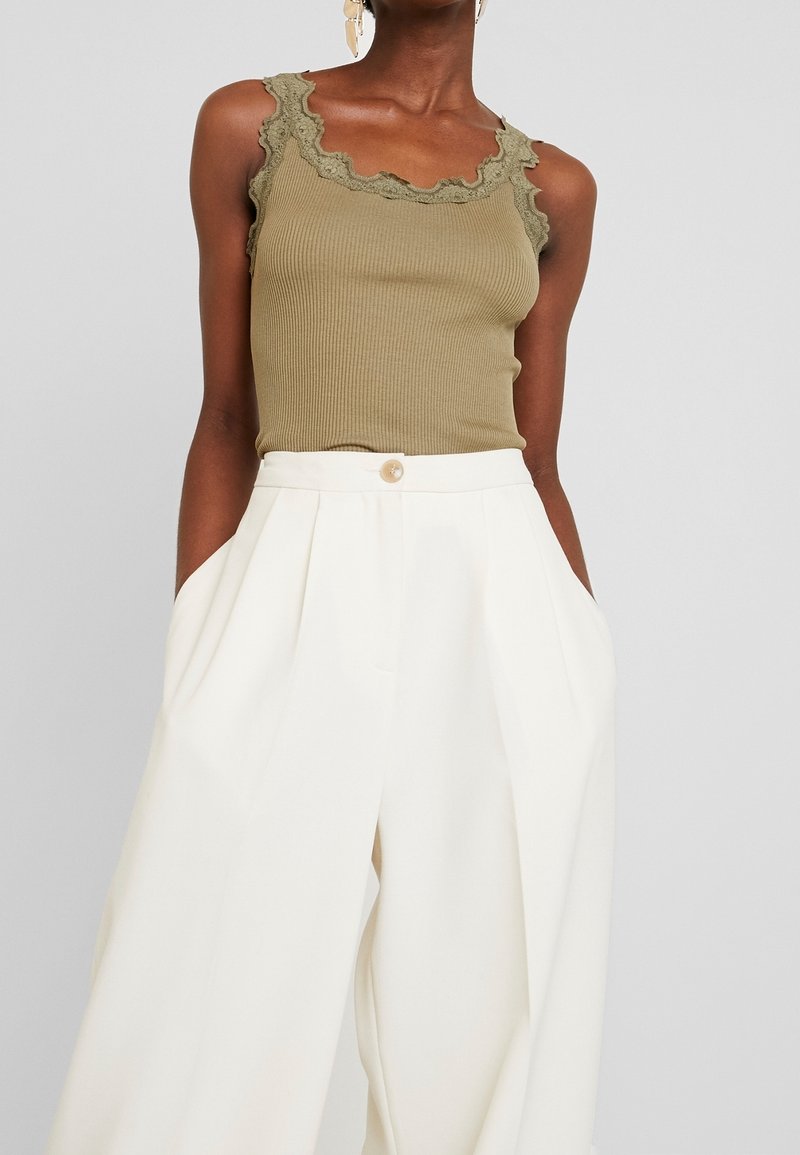 Woman wearing an olive green ribbed tank top with lace trim and high-waisted, wide-leg white pants with pockets and a button closure.