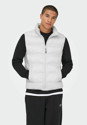 Joven con un chaleco blanco acolchado sobre una camiseta negra de manga larga y pantalones negros, de pie con las manos en los bolsillos del chaleco.