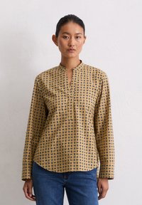 Langarmshirt in een mosterdgele stof met een geometrisch blauw patroon, voorzien van een ingesneden kraag en een ronde zoom.