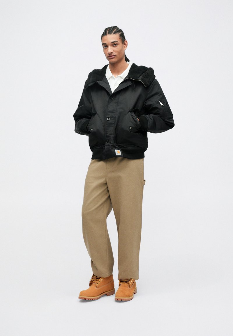 Veste bomber noire avec une capuche en fausse fourrure texturée, devant à boutons, et poches latérales, assortie à un pantalon beige et des bottes de travail marron.