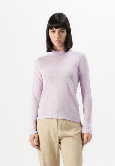 Mujer con cabello oscuro liso que lleva un suéter de cuello alto de manga larga morado claro y pantalones beige texturizados, de pie frente a un fondo blanco liso.