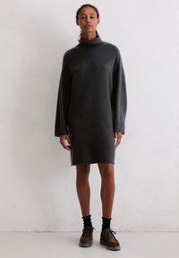 Oversized szary sukienka w kolorze węgla z wysokim kołnierzem, długimi rękawami i ściągaczami. Miękka tekstura, sięgająca do połowy uda. Połączona z brązowymi butami.