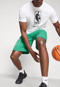 Fehér póló fekete NBA logóval, zöld rövidnadrág fehér felirattal, és fekete sportcipők fehér részletekkel. Játékos egy kosárlabdát tart a kezében.