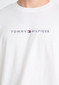 Bijela pamučna majica s natpisom "TOMMY HILFIGER" u mornarskoj boji, s crveno-plavom zastavom koja naglašava slovo 'H'.