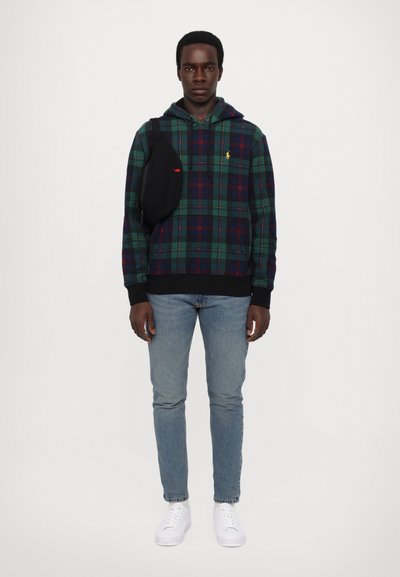 Giovane in piedi, indossa un hoodie a quadri verdi e blu, jeans azzurri chiari, sneakers bianche e una borsa a tracolla nera su uno sfondo bianco.