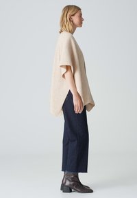 Poncho en tricot beige à manches courtes, doté d'une coupe ample et d'un motif texturé, associé à un jean droit bleu foncé et des bottines.