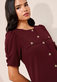 Bluza burgundy cu mâneci bufante, cu decolteu rotund, două buzunare frontale și nasturi decorativi în formă de floare. Material moale cu textură fină.