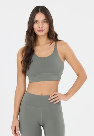 Vrouw met lang donker haar die een bijpassende grijze sportbeha en leggings met hoge taille draagt, staand met één hand op de heup tegen een witte achtergrond.