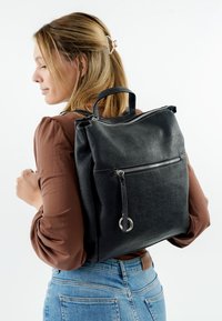 Vaaleahiuksinen nuori nainen ruskeassa paidassa kantaa mustaa teksturoitua nahkareppua yhden olkapään yli valkoista taustaa vasten.