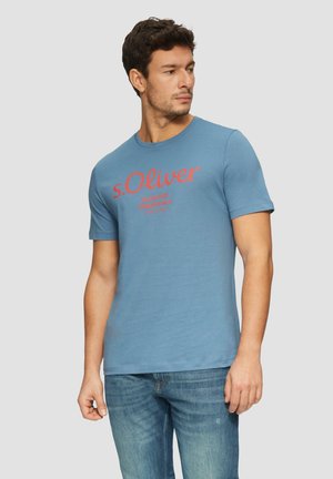Blaues Baumwoll-T-Shirt mit kurzen Ärmeln, rundem Halsausschnitt und rotem Drucklogo. Kombiniert mit hellblauen Jeans.