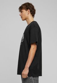 Sort bomulds T-shirt med korte ærmer, afslappet pasform, rund hals, med et grafisk design foran, vist af en person fra siden.