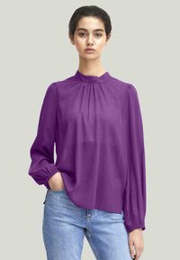 Blusa viola con collo alto, tessuto morbido che cade dolcemente, maniche lunghe con polsini arricciati e una vestibilità rilassata, abbinata a jeans azzurri chiari.