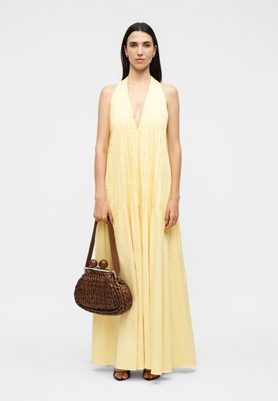 Γυναίκα με μακριά σκούρα μαλλιά που φοράει αμάνικο παλ ανοιχτό κίτρινο μακρύ φόρεμα και κρατάει μία μεγάλη καφέ πλεκτή τσάντα με στρογγυλό κούμπωμα.