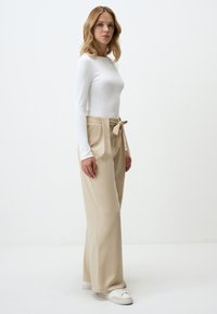 Witte geribbelde top met lange mouwen gecombineerd met hooggesneden beige broeken met een strikceintuur; model draagt witte sneakers. Soepele textuur.