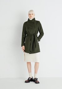 Cappotto verde oliva con cintura, colletto alto e tasche laterali, realizzato in un tessuto morbido. Indossato sopra una gonna in maglia beige chiaro e scarpe scure.
