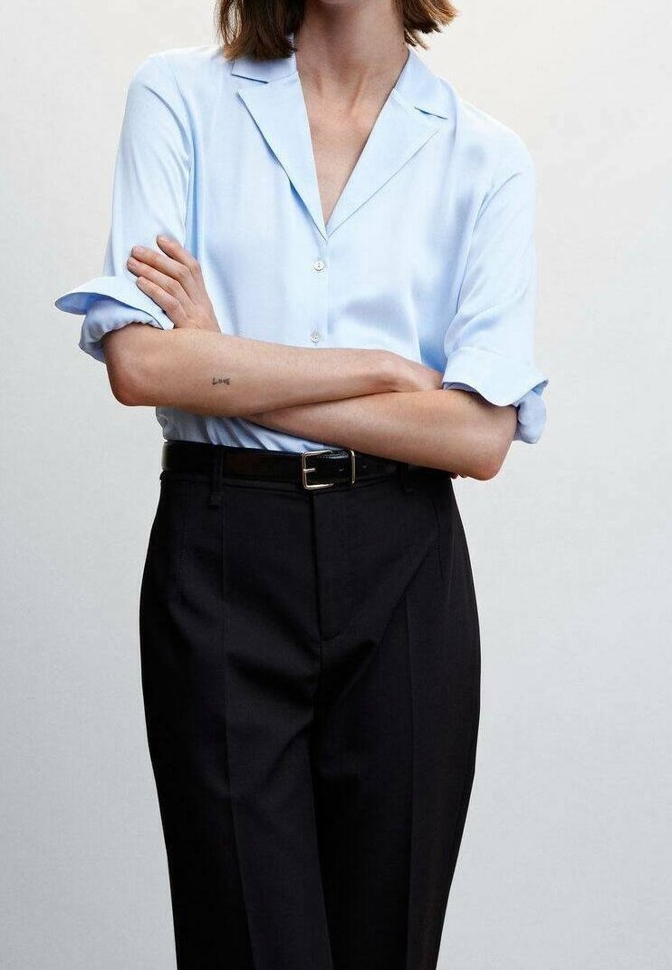 Personne aux bras croisés portant une chemise bleu clair à boutons avec les manches retroussées et un pantalon noir taille haute avec une ceinture noire.