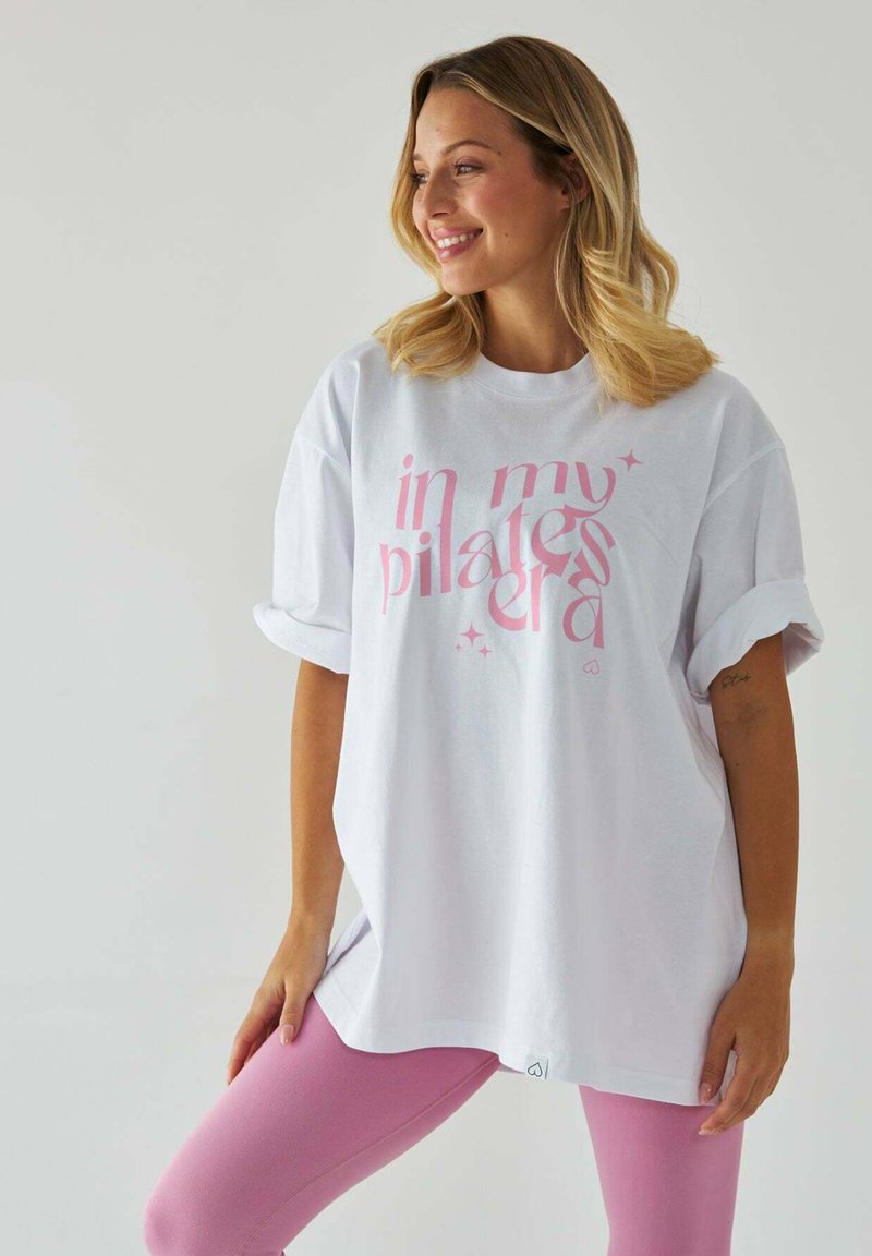 Biały, oversize'owy T-shirt z różowym napisem "w mojej erze pilatesu", w połączeniu z jasnoróżowymi legginsami. Krótkie rękawy i luźny krój.
