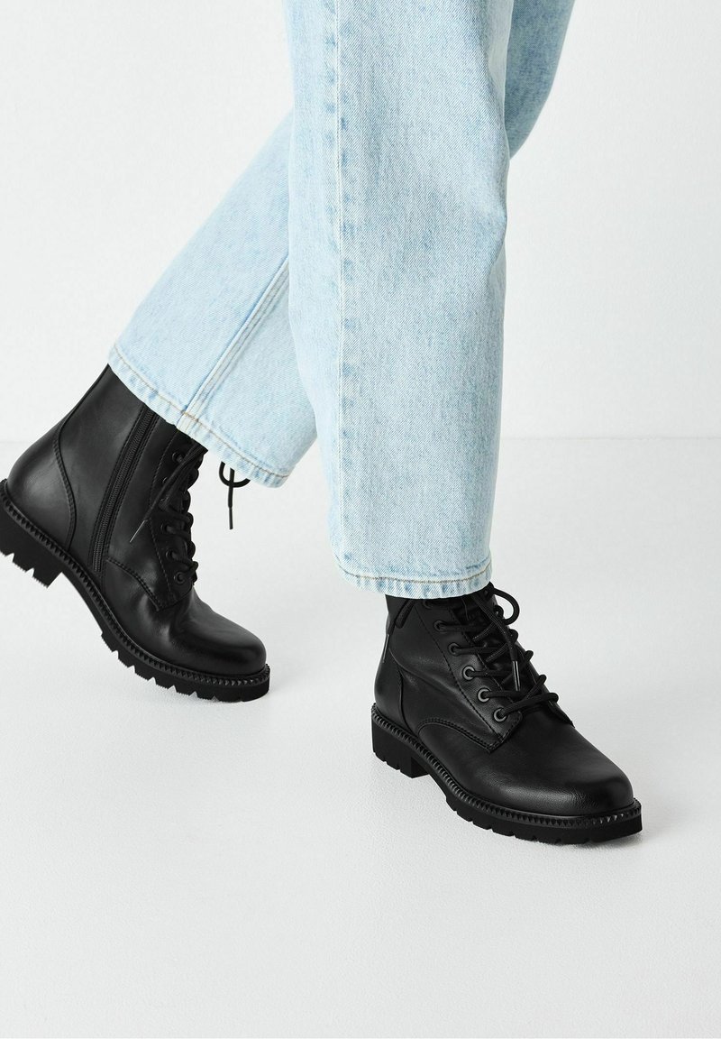 Personne portant un jean bleu clair et des bottines à lacets noires avec des semelles épaisses, debout sur un sol blanc.