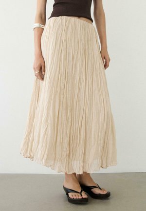 Kobieta ubrana w długą, beżową, pogniecioną spódnicę, czarny bezrękawnik, srebrną bransoletkę, pierścionek oraz czarne sandały na obcasie, stojąca na szarej podłodze.