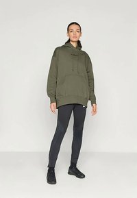 Olivgrüner übergroßer Hoodie mit Kordeln und Kängurutasche, kombiniert mit schwarzen, eng anliegenden Leggings mit strukturierten Einsätzen und Logo-Akzenten.