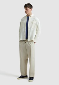 Hellbeige Baumwollhemd, zwei Fronttaschen, offener Kragen; dunkelblaues T-Shirt darunter; beige lockere Hose; dunkelblaue Sneakers mit hellen Sohlen.