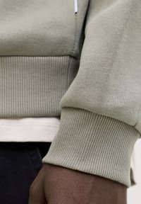 Χέρι σκούρου δέρματος μερικώς ορατό κάτω από γκρι ανοιχτό μανίκι με ρίγες και ζώνη, φορεμένα πάνω από λευκό πουκάμισο και σκούρο παντελόνι.