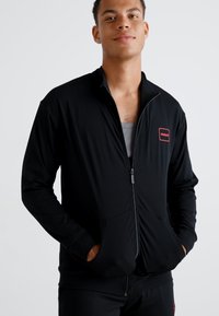 Svart zip-up jakke laget av glatt stoff, med høy krage, lange ermer og en rød logo-patch på brystet. Har to sidelommer.