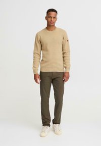 Pulover tricotat de culoare bej deschis, cu guler rotund, mansete și tiv cu nervuri, asortat cu pantaloni verde măsliniu și teniși de culoare crem.