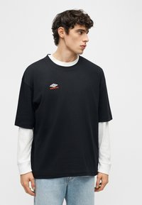 Νεαρός άνδρας με σγουρά μαλλιά, φορώντας μαύρο t-shirt Umbro πάνω από ένα λευκό μακρυμάνικο πουκάμισο και ανοιχτόχρωμο τζιν, κοιτάζει προς το πλάι.