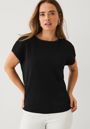 Kobieta o długich włosach, nosząca złote kolczyki, czarny, prążkowany top z krótkim rękawem oraz białe spodnie, pozująca na tle jednolitego, jasnego tła.