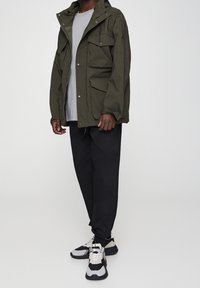 Olijfgroene waterdichte jas met capuchon, meerdere voorzakken en een relaxte pasvorm, gecombineerd met zwarte broek en grijs-zwart sneakers.