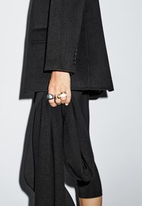 Mano che tiene un maglione nero lavorato a maglia, adornato con anelli spessi d'argento e oro, indossando un blazer nero e leggings neri su uno sfondo neutro.