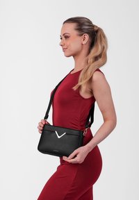 Černá kožená crossbody taška s texturovaným povrchem, stříbrným V-tvarovaným akcentem a nastavitelným popruhem, kterou drží modelka v červených šatech.