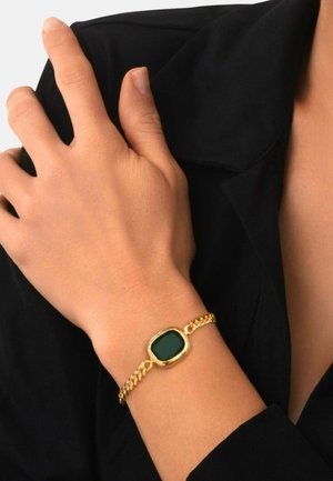 Poignet avec un bracelet chaîne en or orné d'une pierre rectangulaire vert foncé, reposant sur un tissu noir près d'un col plié.