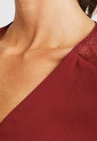 Tissu bordeaux à la texture lisse, agrémenté d'un détail en dentelle à l'épaule. Découpé en forme de V, laissant entrevoir la couleur de la peau sous le tissu.