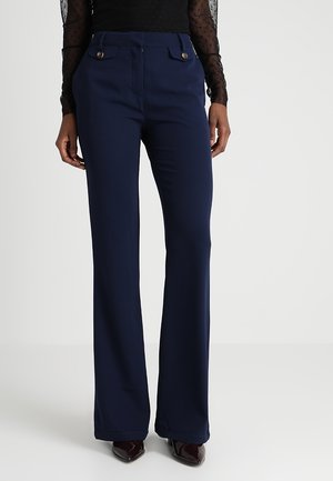 Vrouw draagt marineblauwe broek met hoge taille en met knopen gesloten voorkantzakken, gecombineerd met een doorschijnende zwarte top met lange mouwen en donkere puntige schoenen.