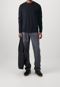 Granatowy sweter z dzianiny, okrągły dekolt, w połączeniu z jasnoszarymi, nieco luźnymi dżinsami. Czarne sneakersy z brązową podeszwą, trzymający czarną torbę.